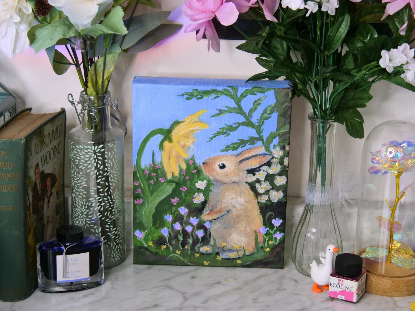 Decorative setup with a painting of a rabbit, flowers, and books on a marble surface.