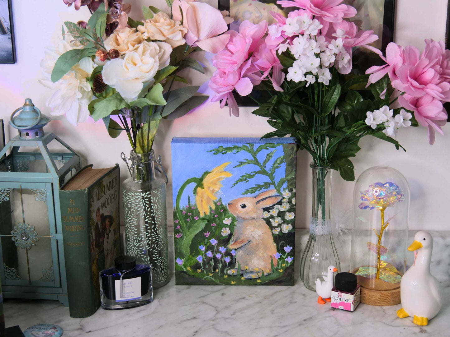 Decorative setup with flowers, a painting of a rabbit, and small figurines on a marble surface.