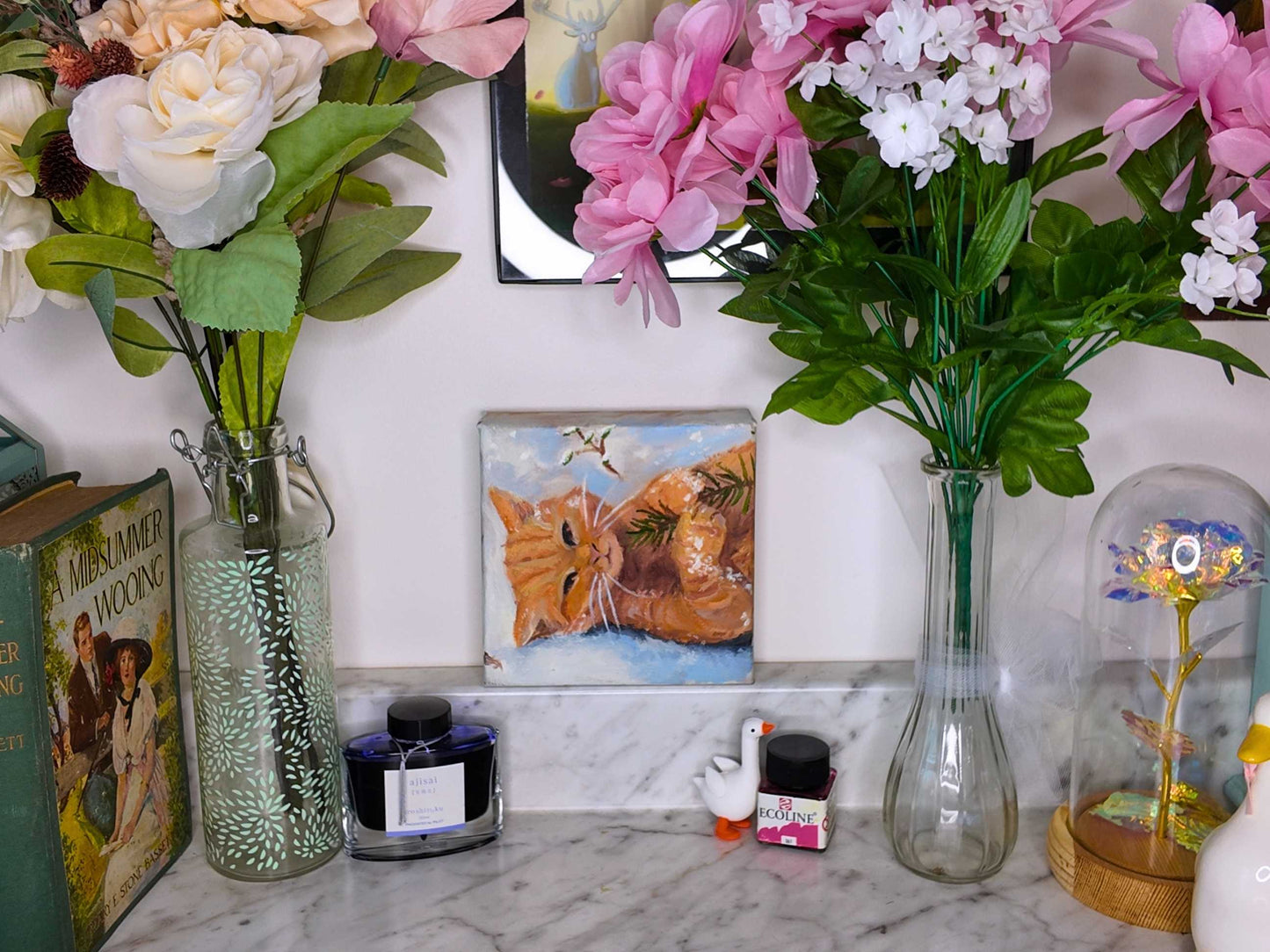 Decorative setup with flowers in vases, a painting of a cat, and books on a marble surface.